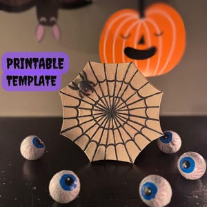 Op de afbeelding: Een Halloween-display met een spinnenweb, een spin en oogdecoraties. Een lachende oranje pompoen en een vleermuis staan op de achtergrond. De woorden "PRINTABLE TEMPLATE" staan in een paarse doos.