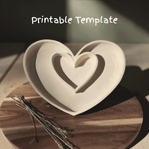 Puede incluir: Un plato de cerámica blanco en forma de corazón con un recorte en forma de corazón más pequeño en el interior. El plato está sobre una superficie de madera.