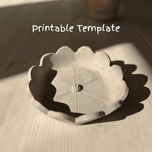 Puede incluir: Una plantilla imprimible para un plato de cerámica con forma de flor con un centro elevado y cinco líneas que irradian desde el centro. El plato es de color gris claro y tiene un borde festoneado.