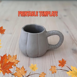 May include: A gray ceramic mug shaped like a pumpkin with a textured handle. The mug has vertical ridges and the words "PRINTABLE TEMPLATE" in orange text. Autumn leaves are scattered around the mug on a light wood surface.