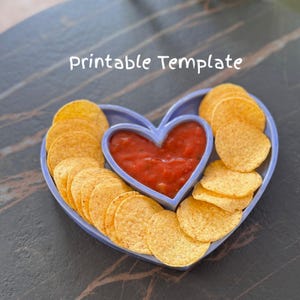 May include: A heart-shaped serving dish with a light blue rim, filled with tortilla chips and red salsa. The centre of the dish is also heart-shaped. The words "Printable Template" are written in white cursive.