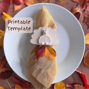 May include: A white ceramic turkey napkin ring adorns a folded tan napkin on a white plate. The napkin ring has a turkey design with a fan-shaped tail. Two colorful fall leaves are tucked under the ring. The words "Printable Template" are in the upper left corner.