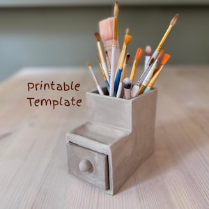 May include: A gray concrete desk organizer with a small drawer and a stepped design. The organizer holds various paint brushes with wooden handles and different colored bristles. The text "Printable Template" is visible on the left.