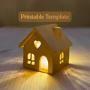 Pode incluir: Uma pequena casa de cerâmica amarela com uma janela em forma de coração e janelas retangulares. A casa tem uma chaminé e o texto "Printable Template" num retângulo castanho por cima. A casa é iluminada por dentro, emitindo um brilho quente.