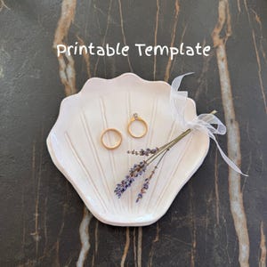 Puede incluir: Un plato blanco en forma de concha con dos anillos de oro y una ramita de lavanda atada con una cinta blanca. Las palabras "Printable Template" están escritas en blanco en la parte superior de la imagen.