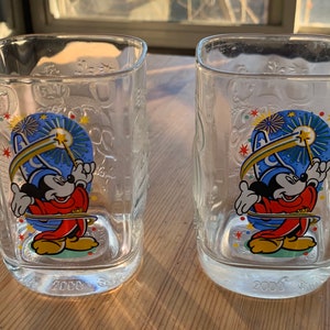 May include: Two clear glass tumblers with a Mickey Mouse design. The tumblers feature Mickey Mouse dressed as a sorcerer, holding a magic wand and a star. The tumblers have a blue, yellow, red, and green design with the year 2000 printed on the bottom.
