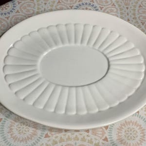 May include: White oval serving platter with a raised, fluted design around the rim and a smooth, oval center. The plate is made of ceramic and is suitable for serving food. The plate is on a patterned surface.