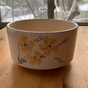 May include: A white ceramic bowl with a yellow floral design. The bowl has a rounded shape and a smooth, white glaze. The flowers are painted in a simple, stylized manner and have a yellow center with brown outlines. The bowl is perfect for serving snacks, salads, or desserts.