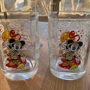 May include: Two clear glass Mickey Mouse cups with a 2000 inscription on the bottom. The cups feature a cartoon image of Mickey Mouse dressed in a safari outfit with a red, yellow, and green color scheme. The cups are decorated with confetti and a red ribbon.