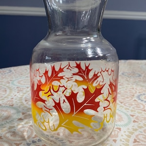 May include: Clear glass pitcher with a wide base and flared top. The pitcher is decorated with colorful autumn leaves in shades of red, orange, and yellow. The leaves are arranged around the bottom of the pitcher.