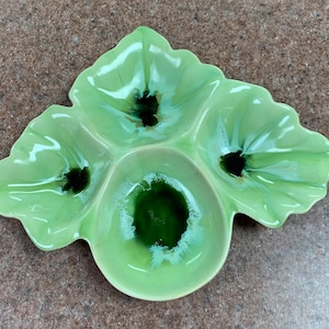 May include: A green ceramic serving dish shaped like a leaf with three sections. The dish has a mottled green glaze with black accents.