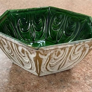 May include: A green ceramic bowl with a geometric shape and a white and gold swirl design on the exterior. The interior of the bowl is a glossy green. The bowl is sitting on a brown surface.