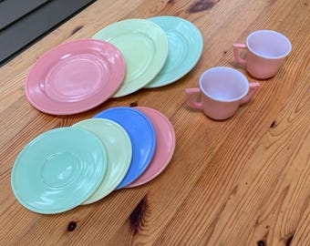 May include: A collection of vintage pastel-colored dishes, including plates and cups. The plates are in shades of pink, green, blue, and yellow. Two pink cups with handles are also present. The dishes are arranged on a wooden surface.