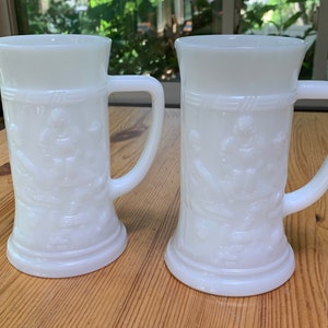 May include: Two white ceramic beer steins with embossed designs. The steins have a handle and a wide base.