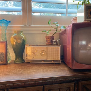 May include: A vintage wooden cabinet with a carved design on the doors. On top of the cabinet, there is a retro clock radio, a green vase, a blue vase, and a television. There is a potted plant on the cabinet and a plant hanging from the window.