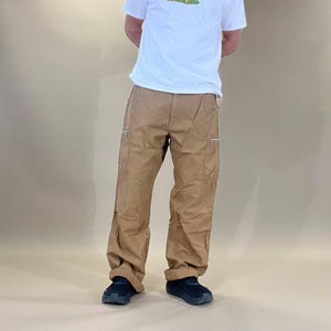 May include: A pair of brown cargo trousers with zipped pockets. The trousers are being worn by a person standing on a light brown surface.