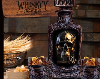 Op de afbeelding: Een donkere, getextureerde glazen whiskykaraf met een schedelafbeelding met gouden vloeistofdruppels en de initialen BH, vergezeld door twee bijpassende glazen met amberkleurige vloeistof en ijs, en een houten bord met de tekst Add Your Name.