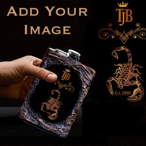以下が含まれることがあります： 黒い革のようなフラスコに、金のサソリのデザインと「TJB Est. 1990」の文字が刻まれています。フラスコは銀色のトップが付いており、手で持たれています。
