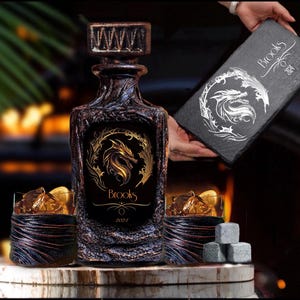 May include: A dark, textured glass decanter with a gold dragon emblem and the text Brooks 2024, accompanied by two matching textured glasses, three grey stone ice cubes, and a black rectangular slate board featuring a white dragon design.