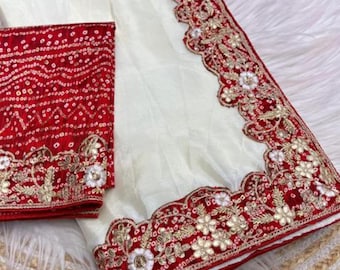 Può includere: Sari color avorio con bordo ricamato rosso e oro. Il sari presenta intricati motivi floreali e geometrici. È incluso anche un pezzo di camicetta rossa abbinato con ricami simili. Il tessuto sembra essere seta o una miscela di seta.
