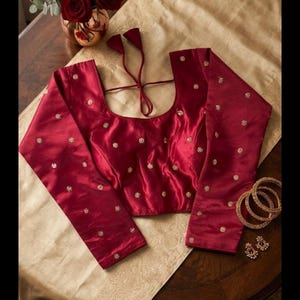 May include: A deep red satin blouse featuring long sleeves and a scoop neckline with tie-up back closures. The garment is adorned with small, scattered gold-toned floral embroidery throughout. Three gold-toned bangles and two matching earrings.