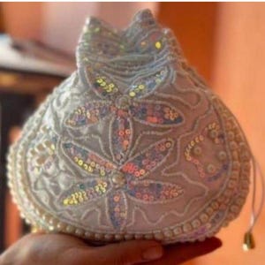 May include: A white drawstring pouch featuring intricate beadwork and iridescent sequins arranged in floral patterns. The bag is embellished with small white pearls along the edges and surface, finished with a simple white cord closure and gold-toned metal tips.