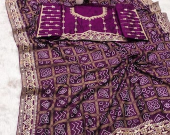 Può includere: Un sari viola con un motivo geometrico dorato e una blusa abbinata. Il sari ha un bordo dorato e la blusa ha ricami dorati intorno allo scollo e alle maniche. Un piccolo vaso di fiori gialli è in cima.