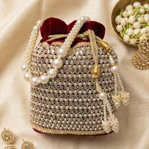 May include: A drawstring pouch bag featuring a deep red velvet top and a body covered in intricate gold embroidery, small mirrors, and white faux pearls. The bag includes a pearl-beaded handle and gold braided drawstrings with pearl tassels.