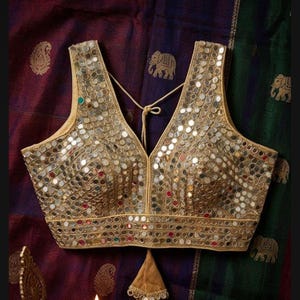 Può includere: Blusa dorata senza maniche, pesantemente impreziosita da piccole paillettes riflettenti. La blusa presenta una scollatura a V e una chiusura con fiocco sul retro. L'indumento è probabilmente per occasioni speciali.