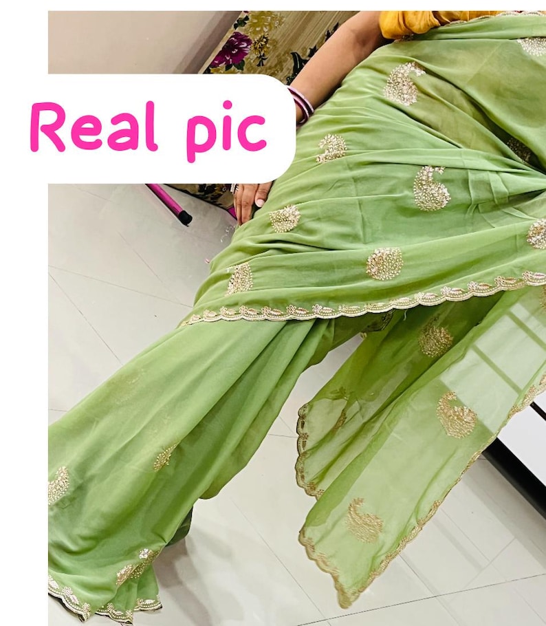 Puede incluir: Un sari verde claro con bordados dorados y un borde festoneado. El sari tiene intrincados detalles dorados y el texto "Real pic" en rosa.