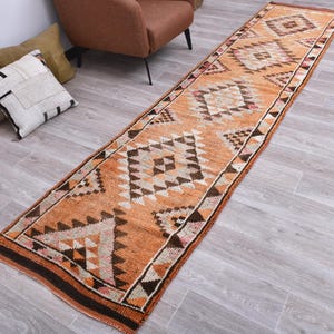 May include: A long, rectangular runner rug featuring a vibrant orange base color with a repeating geometric pattern of diamonds and triangles in shades of dark brown, cream, and white. The rug has a textured, woven appearance with fringed ends.