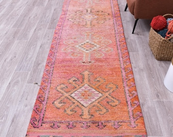 Puede incluir: Una alfombra de pasillo larga y estrecha con un color base naranja vibrante y patrones geométricos de diamantes repetidos en tonos rosa, blanco y marrón oscuro. La alfombra tiene un borde decorativo con un motivo triangular repetido.