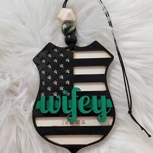 May include: A black and white wooden car air freshener shaped like a police badge with a green "wifey" text and the words "I've got his six."