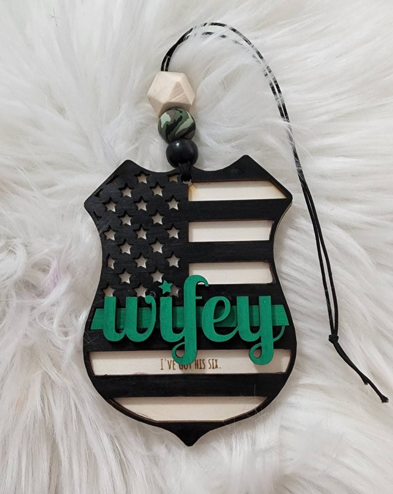 May include: A black and white wooden car air freshener shaped like a police badge with a green "wifey" text and the words "I've got his six."