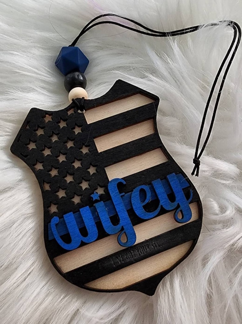 May include: Wooden car air freshener with a blue, white, and black American flag design. The word "wifey" is written in blue letters on the front. The back of the air freshener has the text "I've got your six."