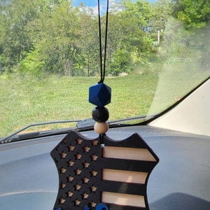 May include: A wooden car charm in the shape of a police badge with an American flag design and the word "wifey" in blue lettering. The charm is hanging from a rearview mirror in a car.