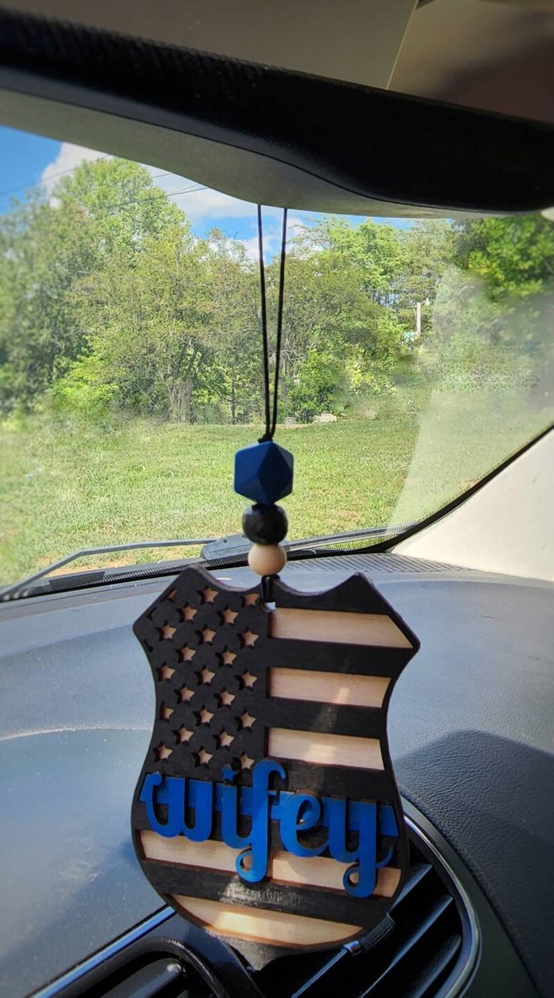 May include: A wooden car charm in the shape of a police badge with an American flag design and the word "wifey" in blue lettering. The charm is hanging from a rearview mirror in a car.