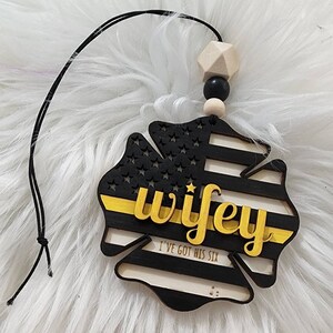 May include: Black and yellow wooden cutout of a fire fighter's helmet with the word "wifey" in gold lettering and the text "I've got his six" below. The cutout is hanging from a black string with wooden beads.