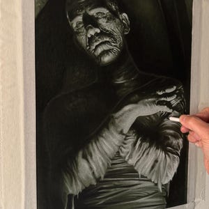 May include: A monochromatic charcoal or graphite drawing on canvas depicting a figure wrapped in textured bandages. The subject has a weathered, aged face with deep-set eyes and a somber expression, rendered in high-contrast black and white tones.
