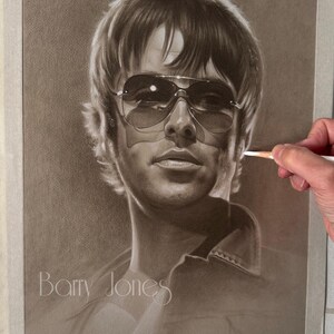 Puede incluir: Dibujo de retrato en sepia de un hombre con gafas de sol. La mano del artista sostiene un lápiz. La obra de arte está pegada a una superficie blanca. El nombre "Barry Jones" está escrito en la obra.