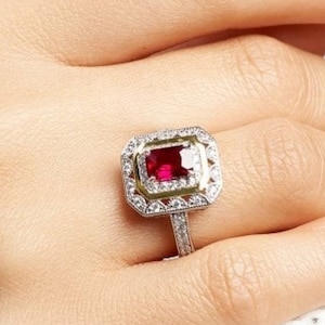 May include: A red gemstone ring with a square-shaped stone surrounded by a halo of smaller diamonds. The ring is set in a silver band with a gold accent.