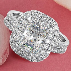 May include: A silver engagement ring featuring a large, square-shaped diamond encircled by a double halo of smaller diamonds. The ring is set on a band adorned with a row of smaller diamonds.