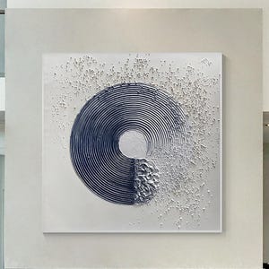 Op de afbeelding: Een vierkant schilderij op canvas met een groot, donkerblauw cirkelvormig motief bestaande uit concentrische lijnen. De rechterkant van de cirkel gaat over in een gestructureerd, verhoogd wit impasto-element, met verspreide witte verfspatten over de witte achtergrond.