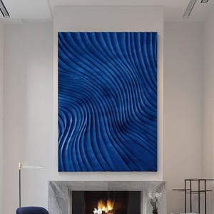 May include: A large rectangular wall art piece featuring deep blue, undulating, wave-like textures. The artwork is mounted above a stone fireplace in a room with two blue armchairs, a blue sofa, a round coffee table, and tall windows.