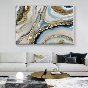 May include: Abstract painting with blue, white, and gold swirls. The painting is framed and hanging on a white wall above a white couch.