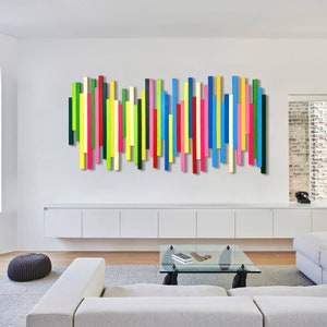 Könnte beinhalten: Eine Wandinstallation bestehend aus zahlreichen vertikalen Holzlatten, die in einer horizontalen Reihe angeordnet sind. Die Latten sind in einem lebendigen Farbspektrum aus Rot, Blau, Gelb, Grün, Pink und Creme lackiert und haben unterschiedliche Längen.