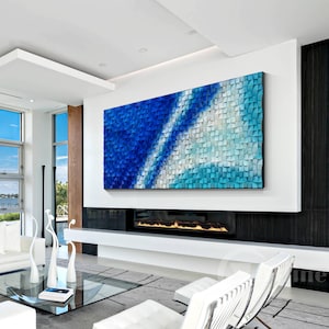 Puede incluir: Una gran pieza de arte de pared abstracta con un esquema de color azul, blanco y turquesa. El arte está hecho de pequeños cuadrados de madera que están dispuestos en un patrón de onda. El arte está colgado en una pared blanca en una sala de estar moderna.