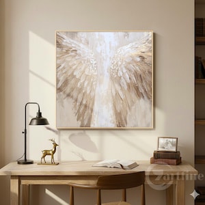 Puede incluir: Un cuadro enmarcado que representa alas de ángel en tonos blancos, beige y dorados. La obra de arte se exhibe sobre un escritorio de madera con una lámpara negra, una figura de ciervo dorada y un libro abierto.