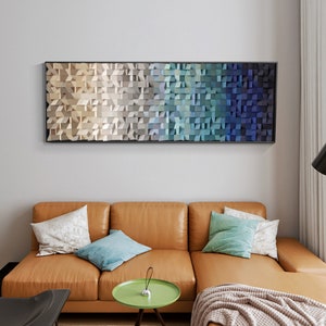 May include: A long, rectangular wall art piece with a geometric design. The design is made up of small, square blocks of wood in shades of beige, blue, and green. The blocks are arranged in a gradient pattern, starting with beige at the left and transitioning to blue at the right.