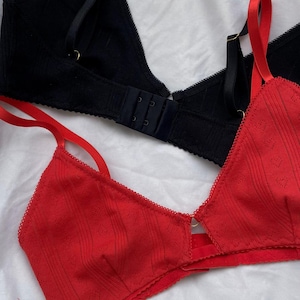 Puede incluir: Un bralette rojo con una sutil textura acanalada vertical y pequeños patrones de corazones, con tirantes rojos finos ajustables y un borde festoneado. Encima hay un bralette negro con una textura acanalada similar y tirantes negros.
